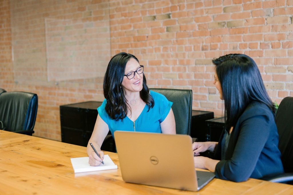 Two business women talking at a wooden conference table in a brick office.