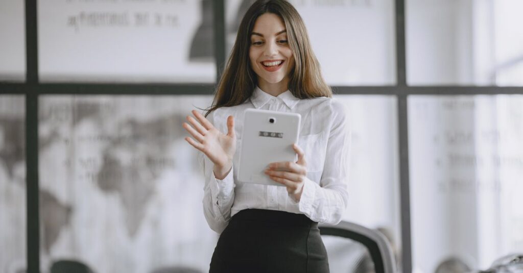 A smiling brunette woman in a professional office setting using a digital tablet for work.