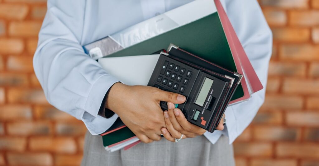 Close-up of a person holding a calculator and folders against a brick wall background.