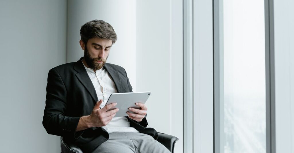 A man in formal attire working on a tablet while seated by a large office window.