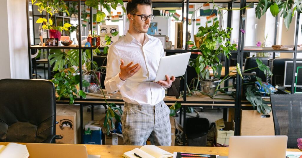Professional man having a video call in a green indoor office space with multiple laptops and potted plants.
