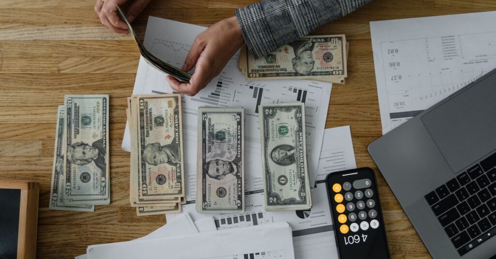 Person counting cash with a calculator and documents on a wooden desk, representing financial planning.