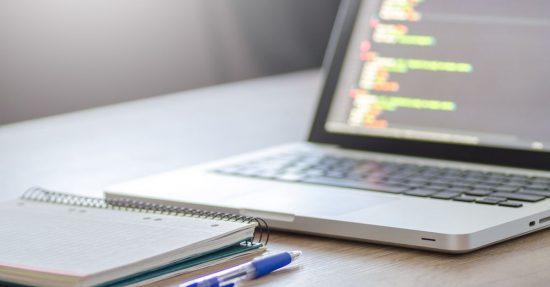 Open laptop with programming code on screen next to a notebook and pen on a desk.