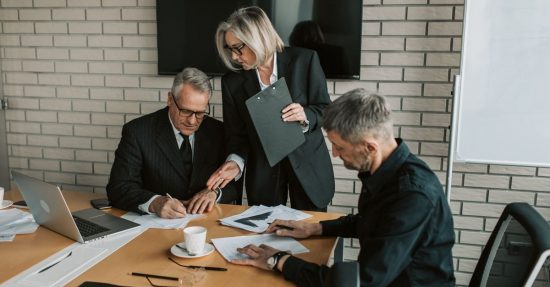 Three mature professionals in a business meeting discussing and signing documents in an office setting.